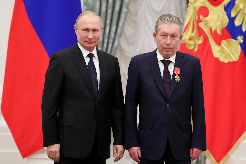two men in suits stand side by side in front of a russian flag