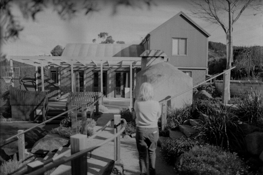 A woman walks towards a house with her back to the camera. 