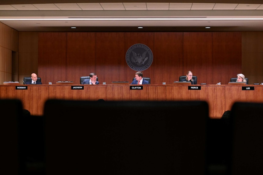 Five people sit at wooden desk in a conference room, with their names displayed underneath.