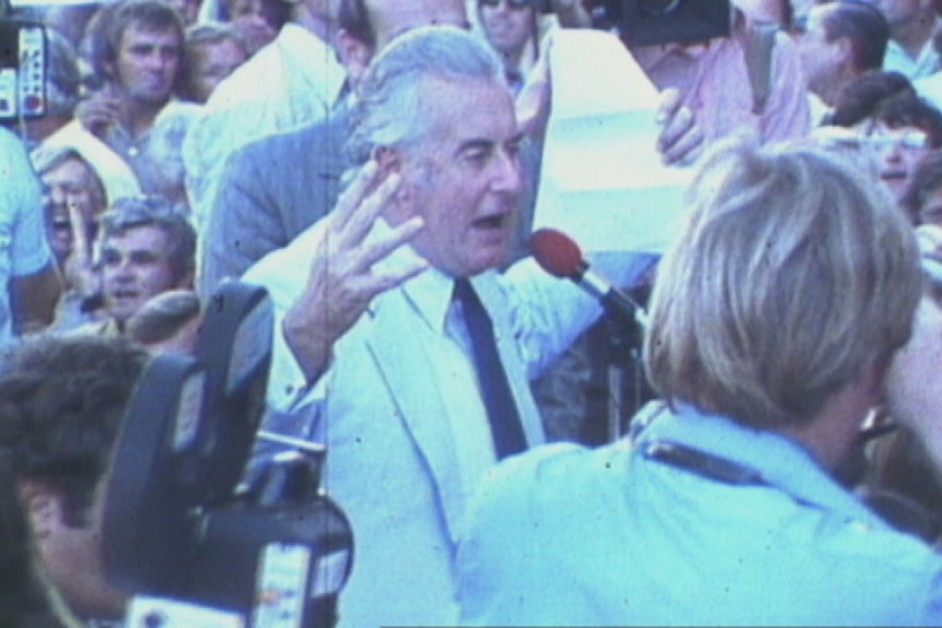 Gough Whitlam, in light suit and dark ties, hold out hands as he speaks into a microphone