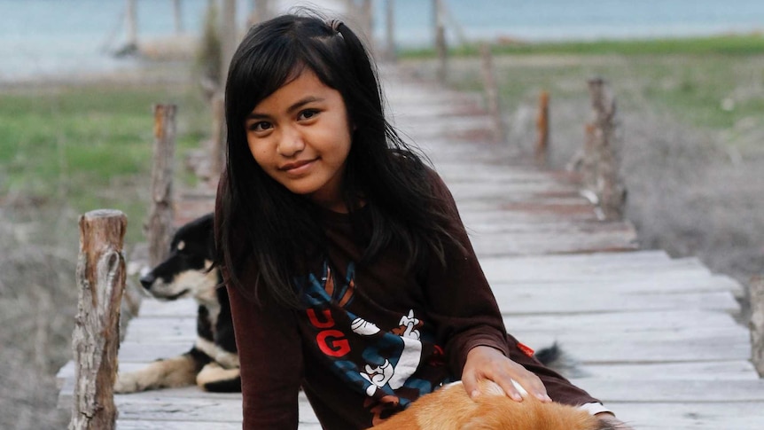 A young girl sits at the foot of a pier in Poso, Indonesia, patting a dog.