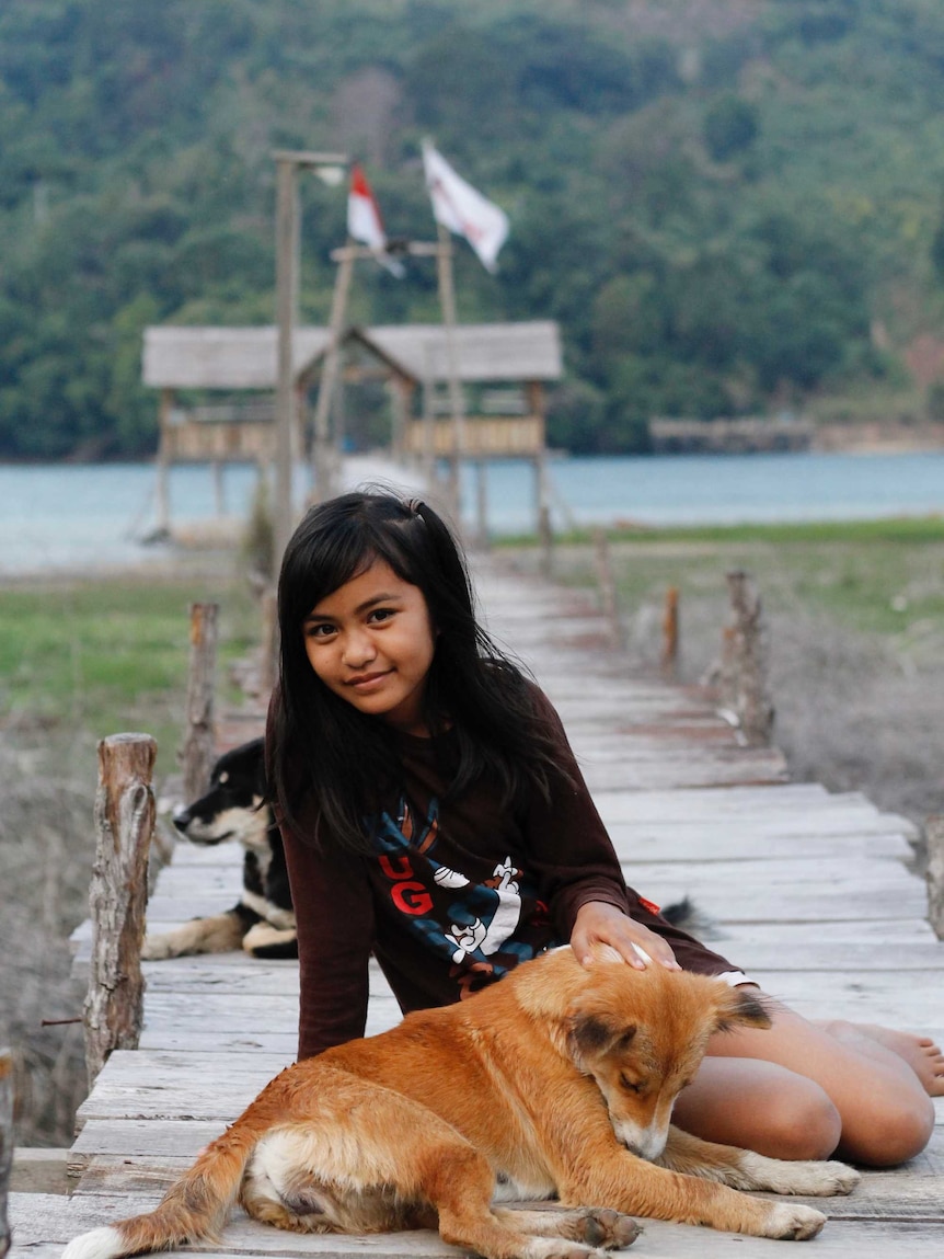 A young girl sits at the foot of a pier in Poso, Indonesia, patting a dog.