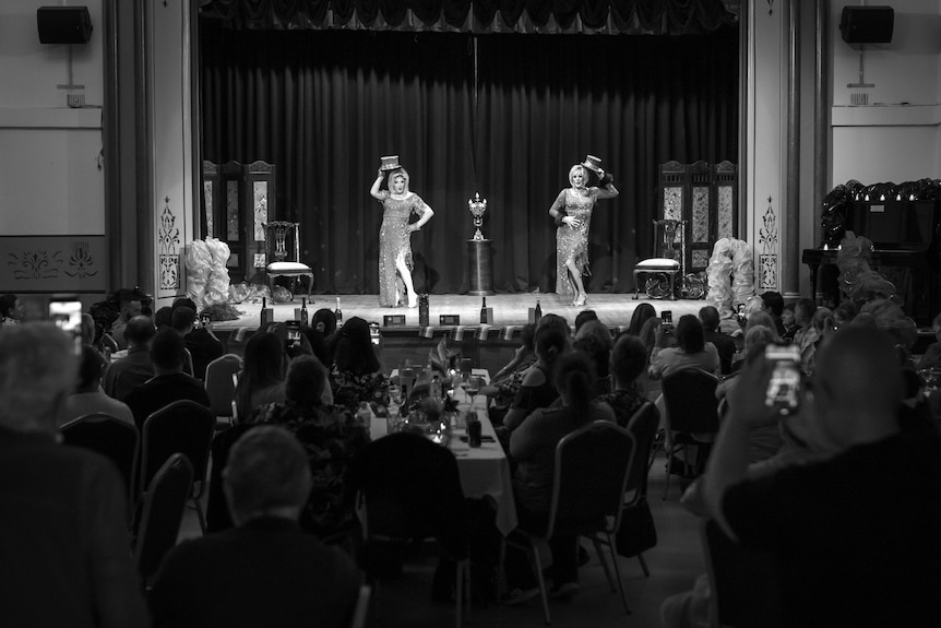 A black and white photo of two drag queens perform on stage in front of an audience. 
