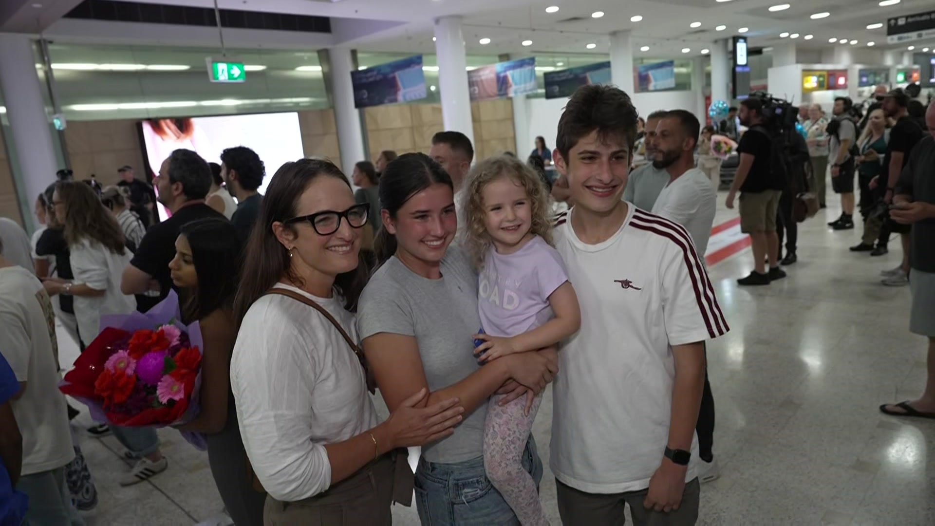 Una madre, dos adolescentes y una niña sonríen ampliamente en una sala abarrotada de aeropuerto.