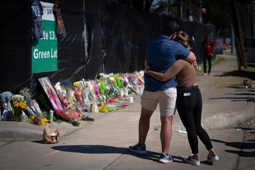 A couple hug while look at flowers laid out along a concert venue fence.