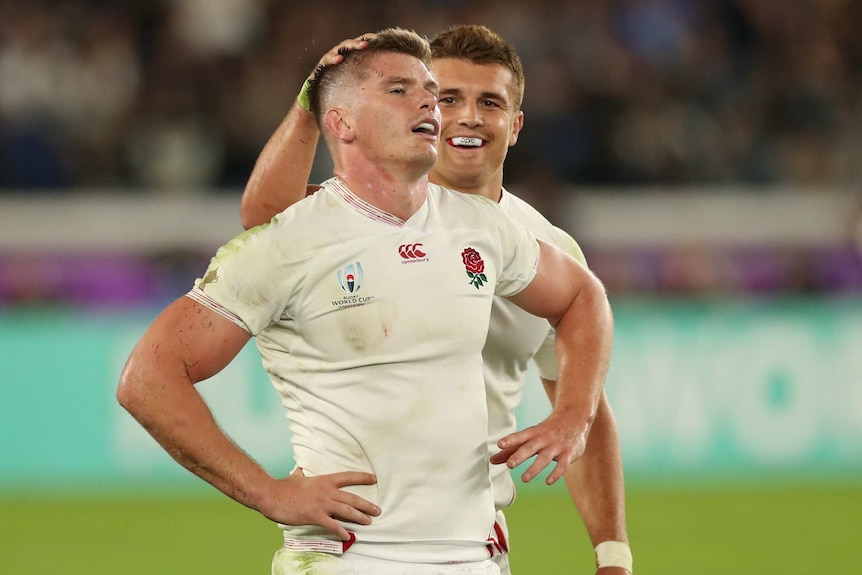 An England male rugby union player stands with his hands on his hips as a teammate stands behind and pats his head.