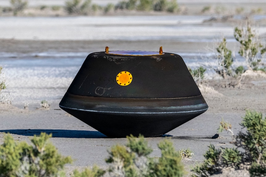 A dark-grey capsule with a yellow port sits on the ground in the desert.