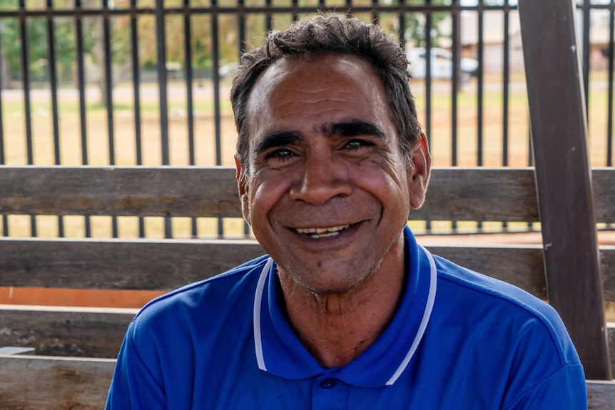 Close up of a middle-aged Aboriginal man, wearing a blue polo shirt, sitting on a bench.