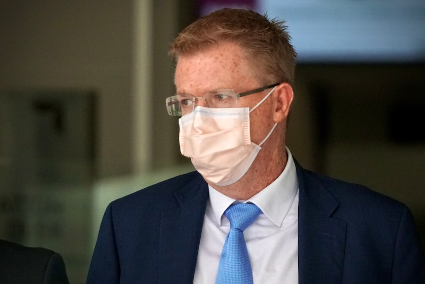 A head and shoulders shot of Kempton Cowan wearing a face mask, suit and tie and spectacles, looking to his right outside court.