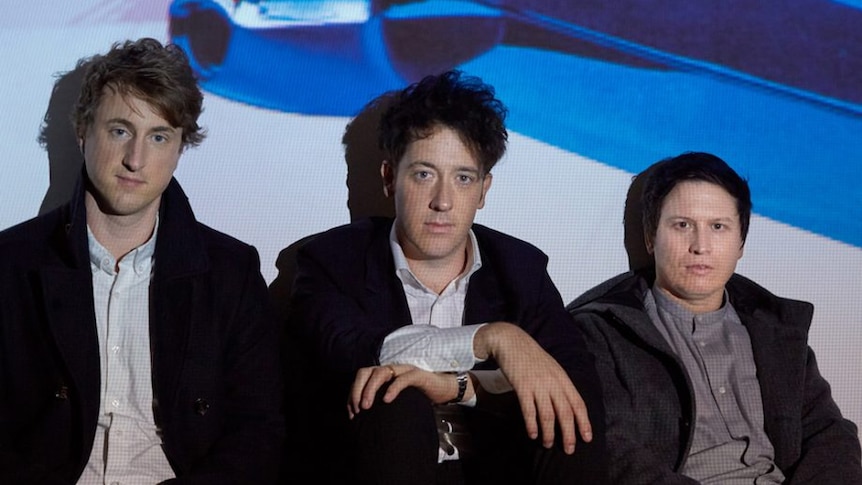 A 2017 press shot of The Wombats sitting in front of a projection of a cadillac