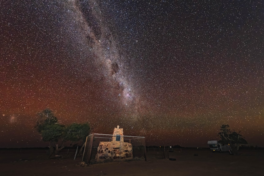 A three tiered memorial cairn sits behind a fence under the bright lights of the Milky Way.