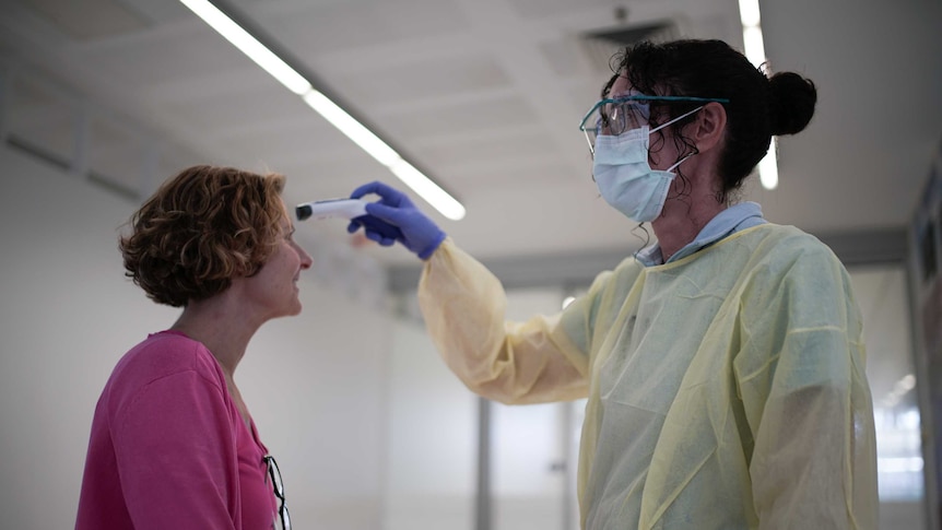 A woman wearing a mask and gown points a thermometer on a woman's forehead