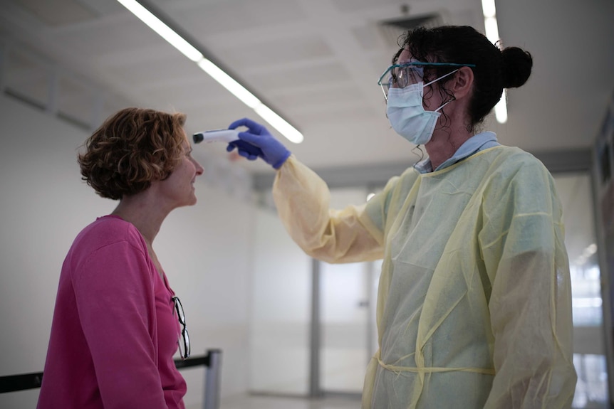 A woman wearing a mask and gown points a thermometer on a woman's forehead