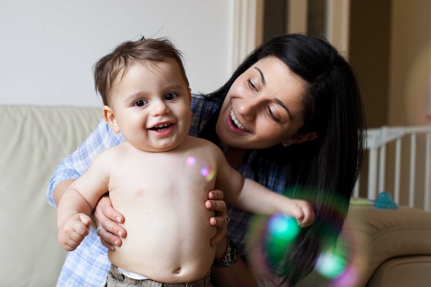A woman holds her toddler son while looking down at him and bubbles float in foreground