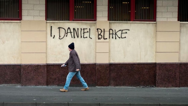 Daniel walks along the street and painted on the wall behind him is I, Daniel Blake, 2016.