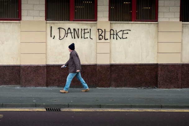 Daniel walks along the street and painted on the wall behind him is I, Daniel Blake, 2016.