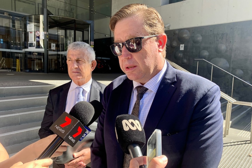 A man in sunglasses speaking into media microphones 