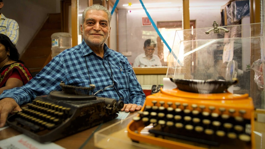 An older man in a buttoned shirt sits, relaxed, with his arm stretched next to one of two old typewriters on a table.