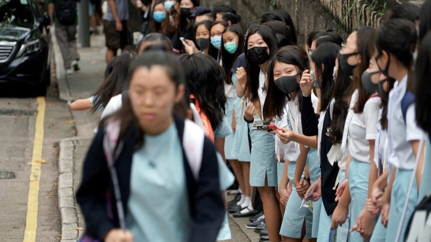 Girls in face masks are lined up shoulder to shoulder at a chain fence.