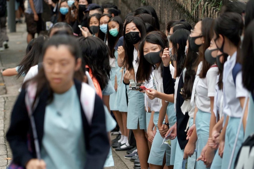 Girls in face masks are lined up shoulder to shoulder at a chain fence.