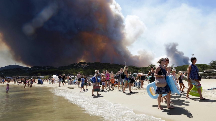 Sunbathers are being evacuated from a beach as plumes of smoke rise behind.