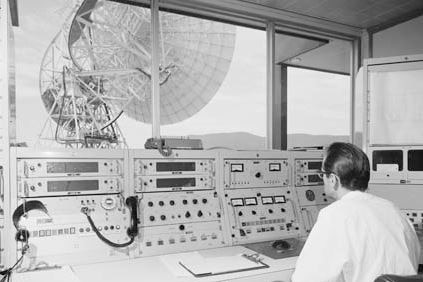 Black and white image of man sitting at instrument panel with view of large radio telescope through window