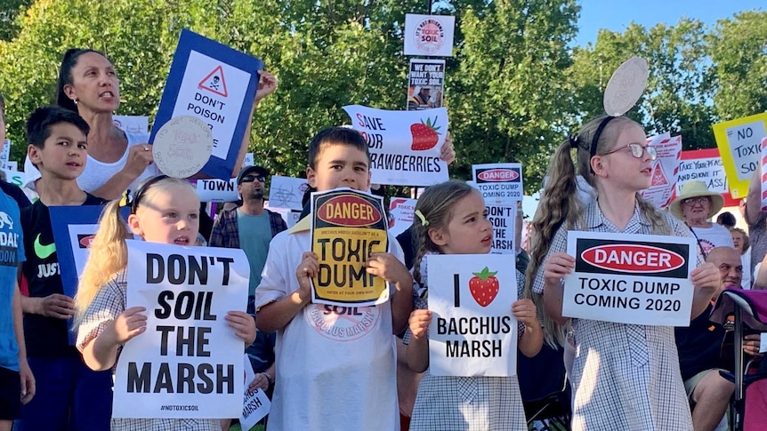 A group of children and an adult hold up signs protesting against a toxic dump.