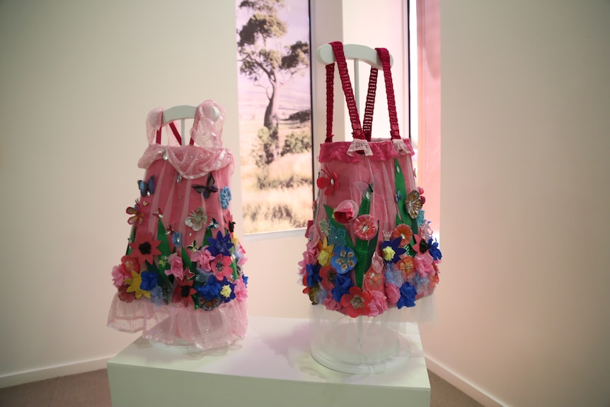 Two pink dresses, one with ruffles, another with straps made out of newspaper in an art gallery. Dresses have colourful flowers.