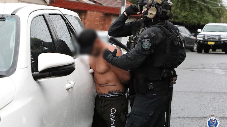 A topless man is arrested by police officers next to a car