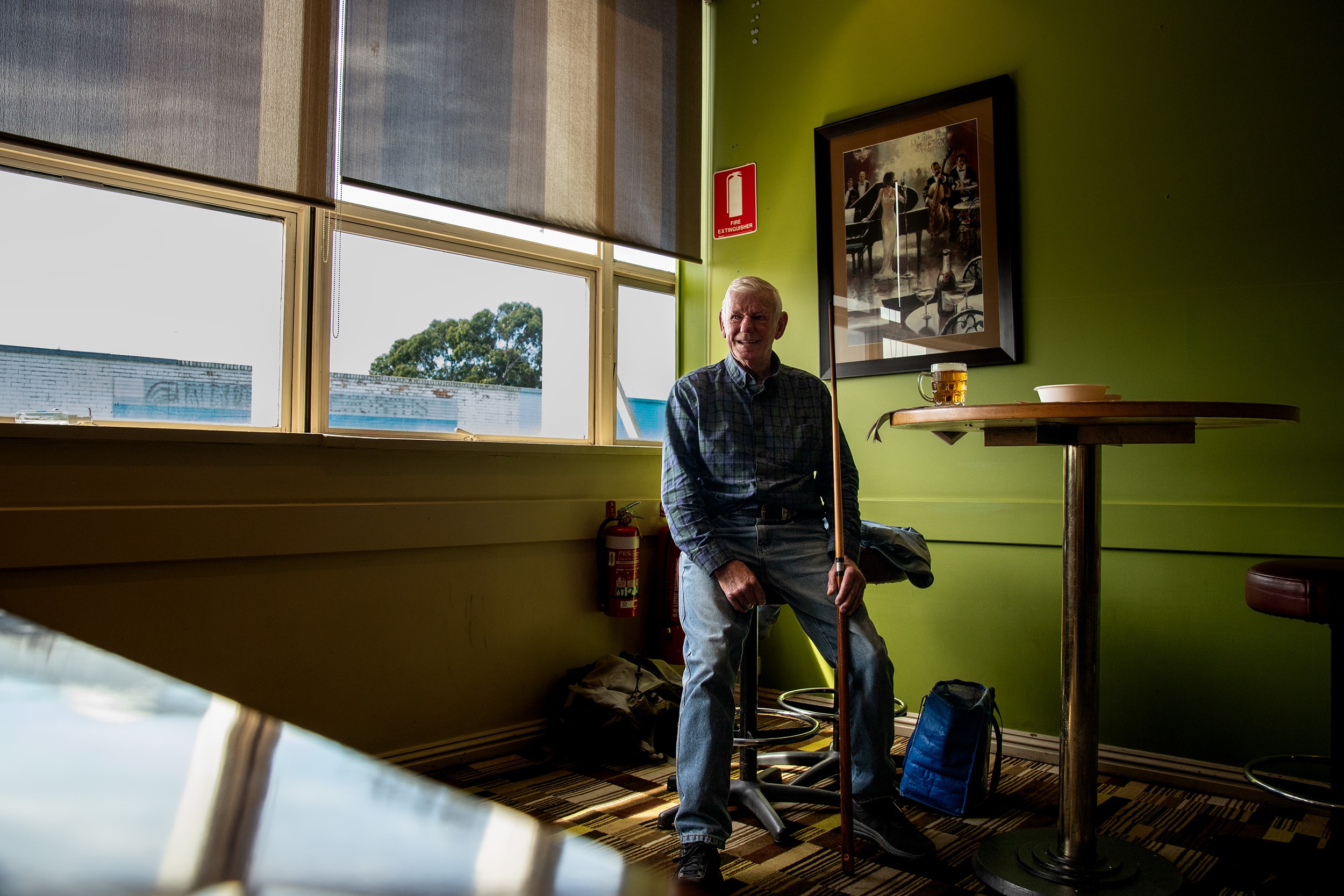 Older man sits at a table with a glass of beer in front of him, holding a pool cue, a bright green wall with an old photo behind