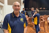 A middle aged man in a sports club polo short stands in a town hall.