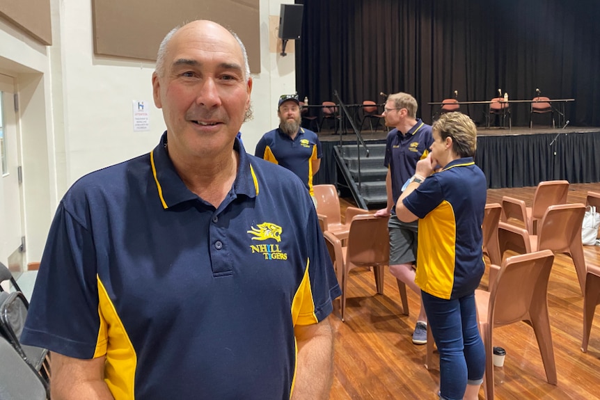 A middle aged man in a sports club polo short stands in a town hall.