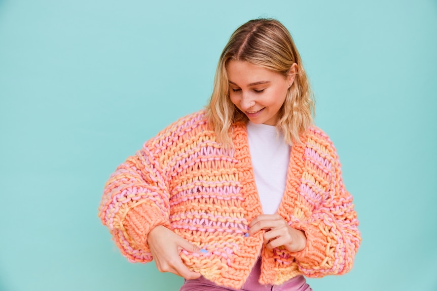 Jaime Dorfman smiles as she looks down and holds the bottom half of an orange and pink chunky knit cardigan she knit.