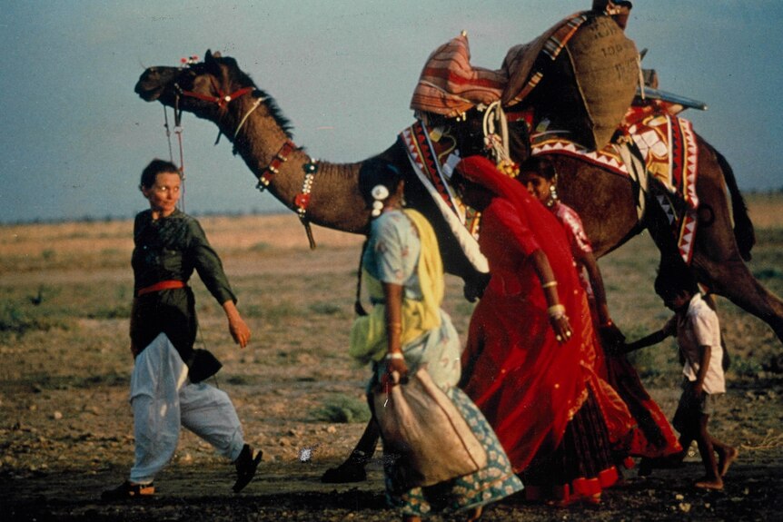 An archive photo of a young Robyn Davidson leading a camel, looking back at three women dressed in colourful saris. 