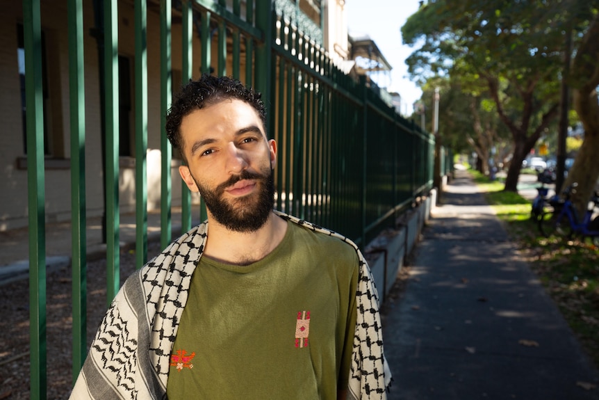 Man with dark hair and beard wearing olive green t-shirt and keffiyeh, standing on street. 