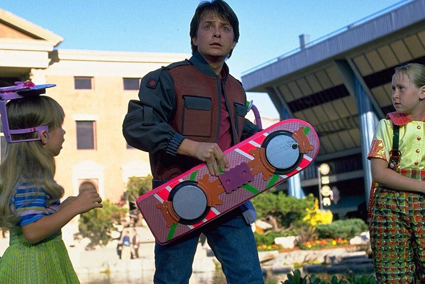 Michael J Fox hold a hoverboard, flanked by two kids.