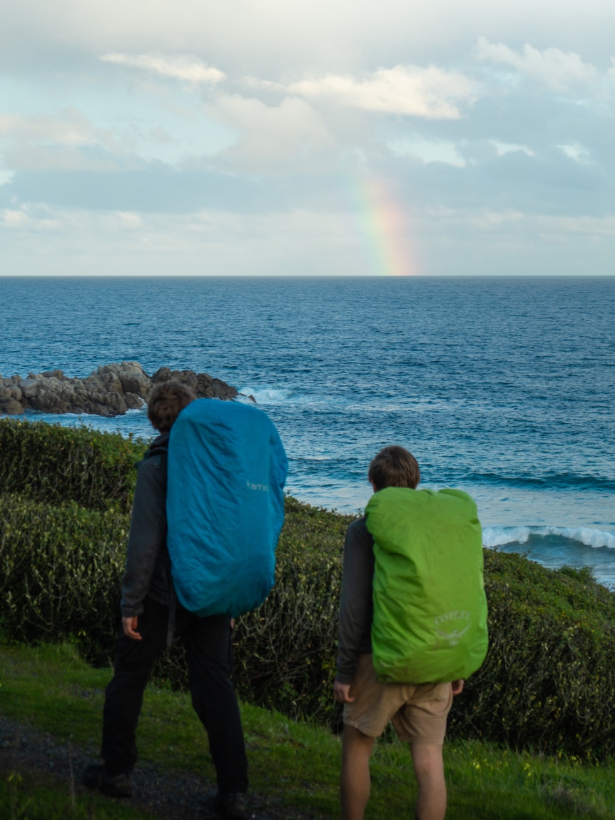 Two boys with camping packs on their backs in front of a cliff with water and a rainbow above