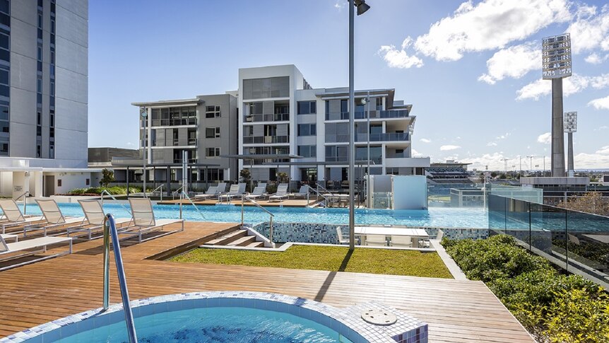 A fourth floor pool deck at the Queens Riverside development in Perth under sunshine with WACA light towers in the distance.