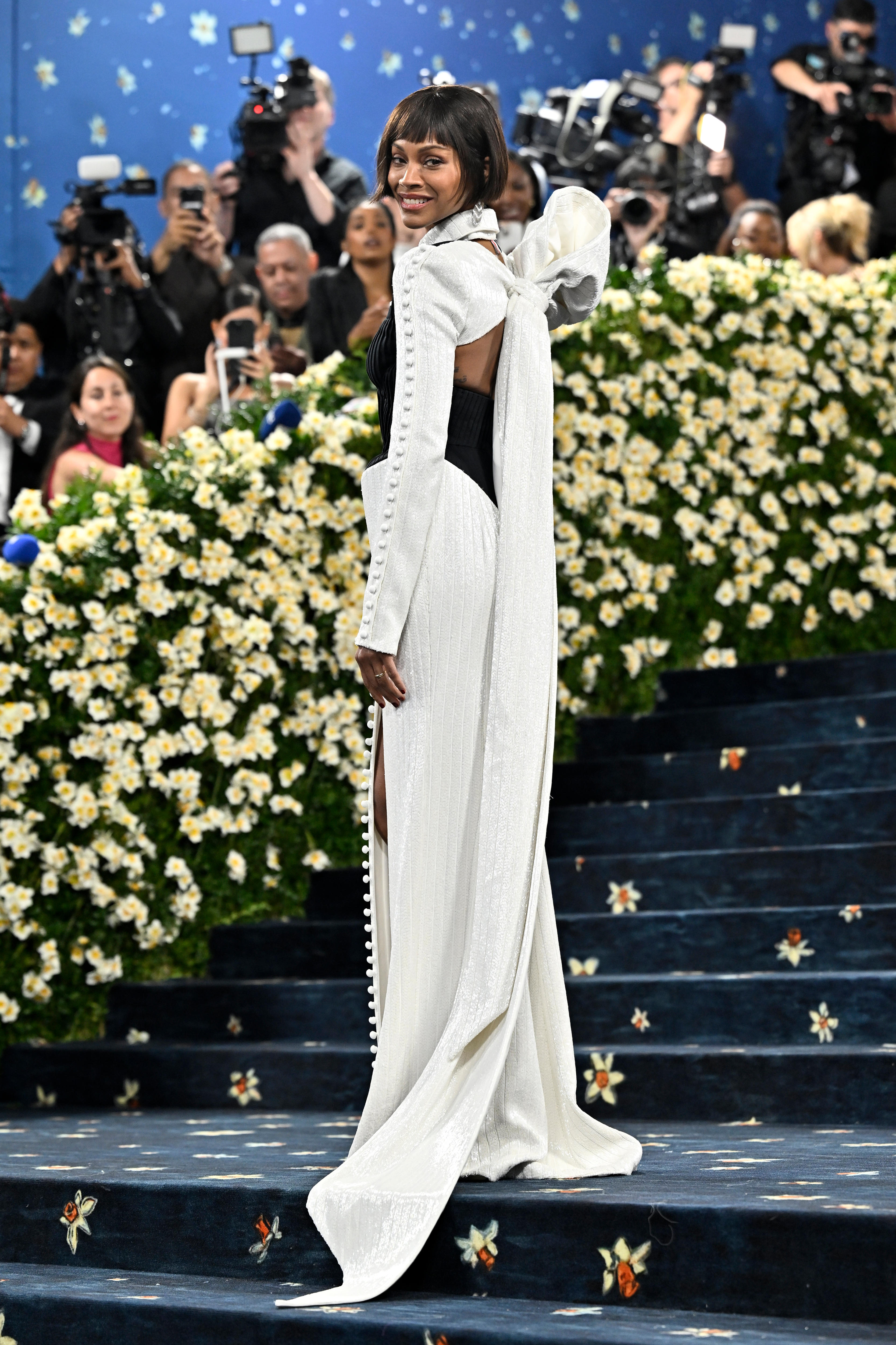 Zoe Saldana wearing a long white gown with long sleeves with buttons all up the side and big bow down her back