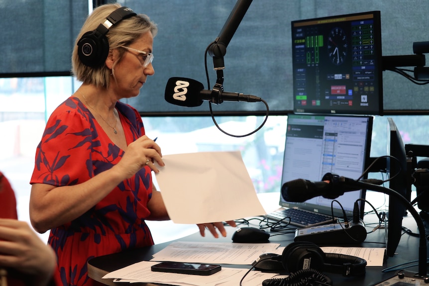 Loretta Ryan wears red printed shirt, headphones, stan behind an ABC radio mic as she turns over a sheet of paper while on air.