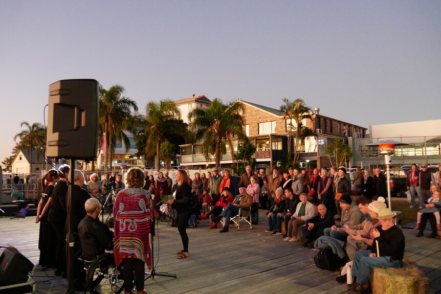 Crowd of people watch performance with buildings and palm trees in the background 