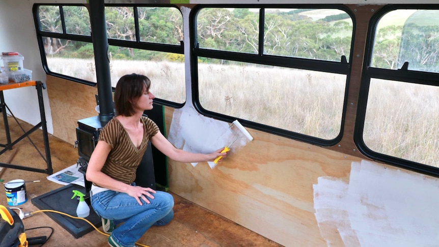 A woman uses a roller to paint the interior of a bus white