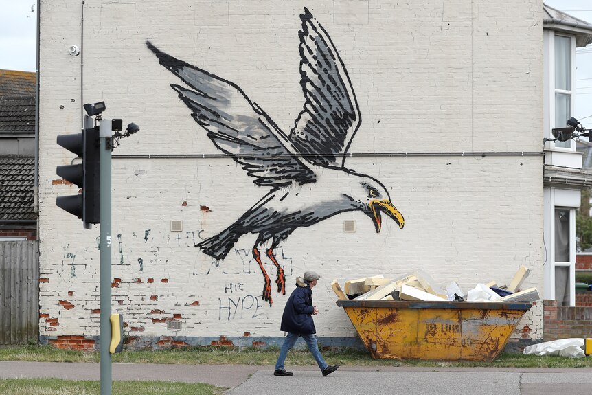 a person walks past a wall where a seagull is painted on it by banksy appearing to swoop toward a skip