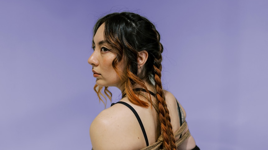 Jaguar Jonze, a Taiwanese Australian woman with braided brown and copper hair stands bound by rope, looking over her shoulder