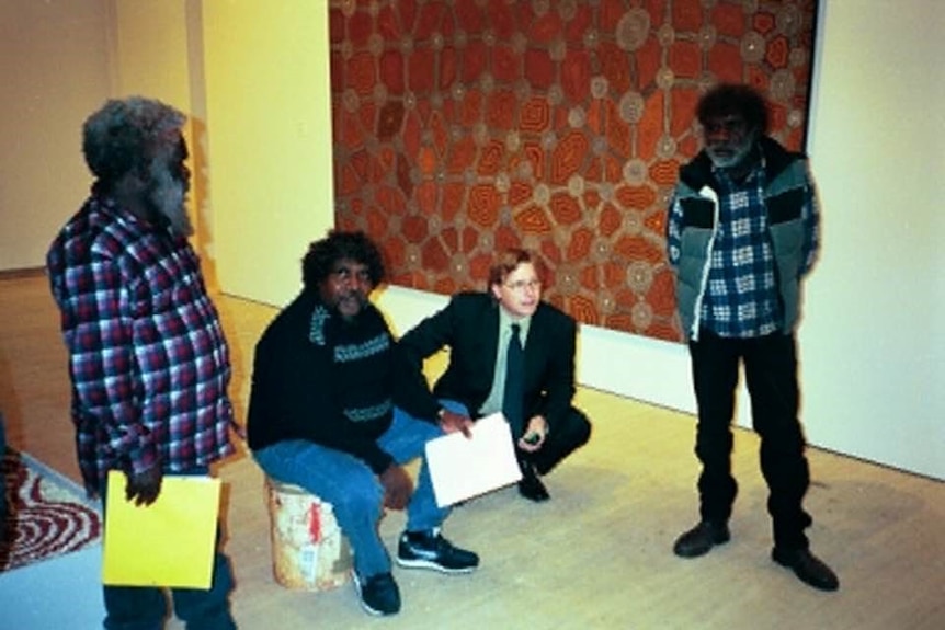 Two older Indigenous men stand, one younger sits on couch, caucasian man in suit crouches on floor, a large artwork on wall.