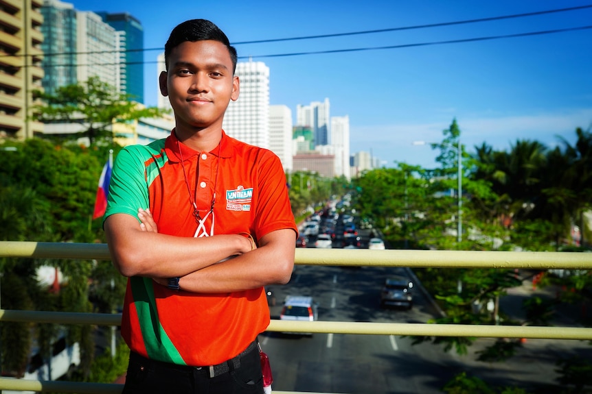 A young man in a red polo shirt stands with his arms crossed in front of a bridge looking out on Manila