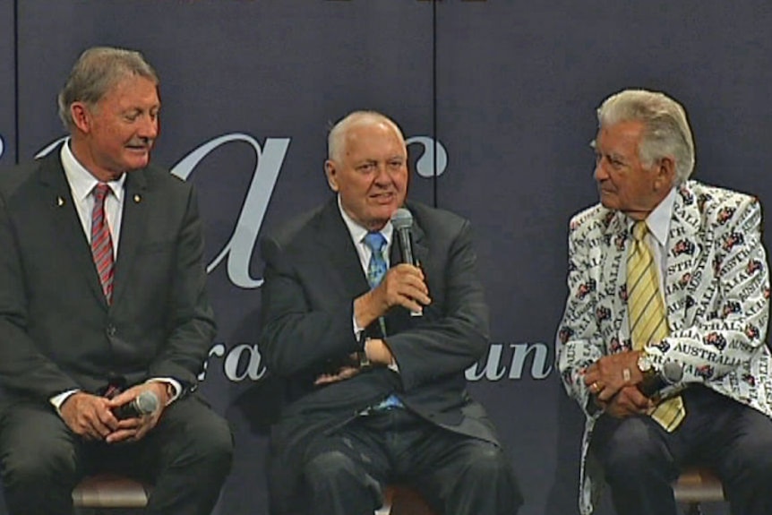 Three men in suits, one in a bold white jacket, sit on a stage
