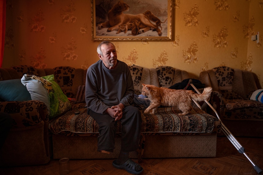 Oleksandr Rabenko sits on a coach next to his orange cat and his crutches