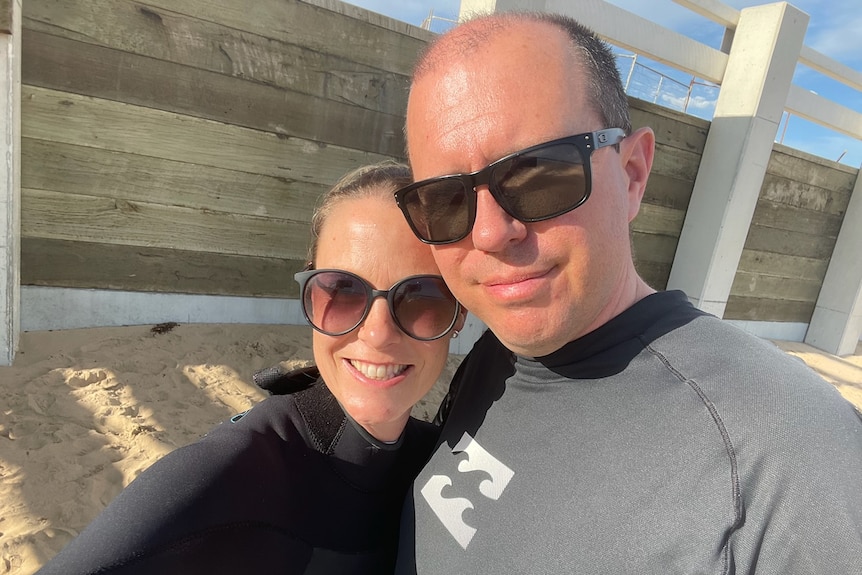 Woman and man in wet suits and sunglasses smile at the camera.