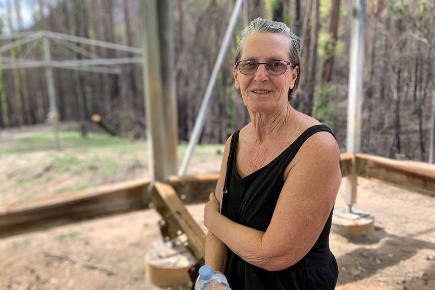 Joan Henderson sits down, holding a drink bottle with burnt bushland in the background.